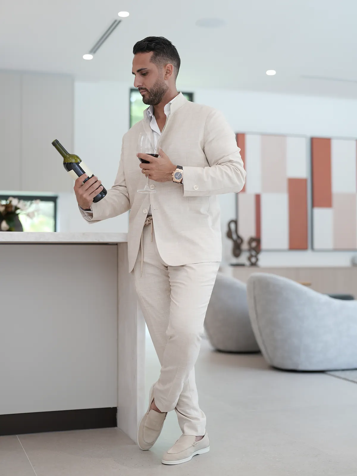 Milano: the beige lapel-less jacket and drawstring trousers, standing at a kitchen counter with a glass of red wine